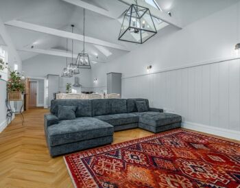 Spacious living room with a dark gray sectional sofa on a red patterned area rug, white paneled walls, and exposed white ceiling beams with geometric pendant lights above.