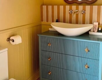 Bathroom featuring yellow beadboard walls, striped wallpaper, and a teal ribbed dresser vanity with brass star handles and a white oval vessel sink.